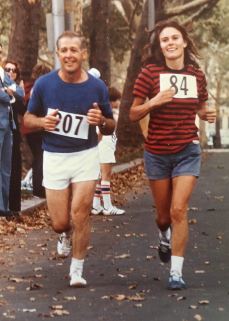 2 runners featured on a fall tree-shaded path flanked by pedestrians.